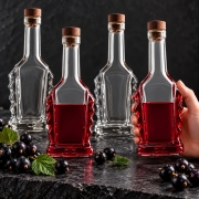 Butelka Bukareszt 250 ml + korek – cztery butelki Bukareszt na ciemnym kamieniu, dwie z czerwonym płynem, jedna w dłoni, czarne porzeczki